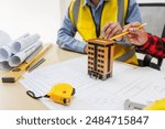 Two men, including a male Asian construction contractor engineer, discuss structural plans and engineering concrete columns and beams of a new house at a desk with a building tower model.