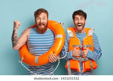 Two men lifeguards use lifeline, wear special orange vest, look happily at camera, saved life of swimmer at beach, does their duties, keep beach safe in summertime period. Life savers watch sea