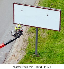 Two Men In A Lift Basket Change Image On A Billboard Near A Road