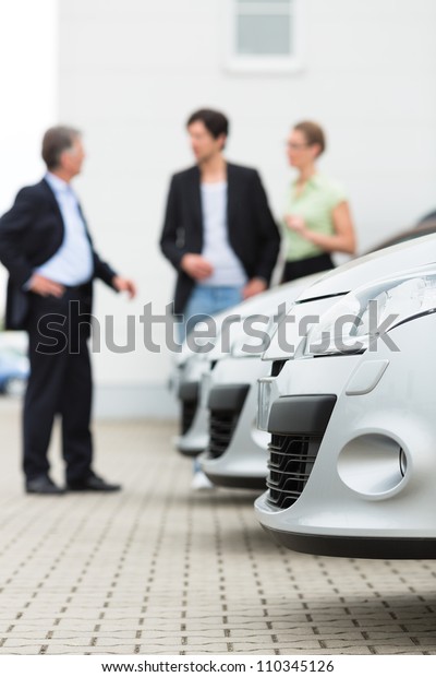 Two men man and one\
woman stand behind a row of silver-metallic cars on a parking place\
on yard of dealer