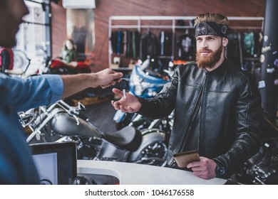 Two Men In Motorcycle Shop. Customer And Salesman. Shop Assistant Is Helping Bearded Man In Choosing New Vehicle, Motorcycle Accessories And Other Biker Stuff. Giving Keys To New Owner.