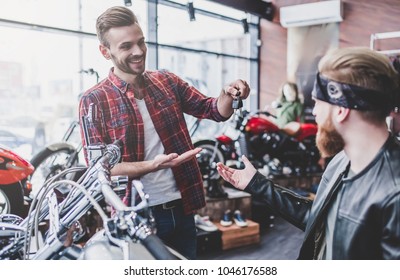 Two Men In Motorcycle Shop. Customer And Salesman. Shop Assistant Is Helping Bearded Man In Choosing New Vehicle, Motorcycle Accessories And Other Biker Stuff. Giving Keys To New Owner.