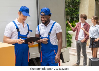 Two Men Movers Worker In Blue Uniform Checking Lists On Clipboard While Unloading Cardboard Boxes From Truck.Young Man And Woman Homeowner Moving To New House.