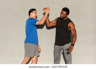 Two men, one Asian and one Black, high-five. The men are smiling, wearing athletic workout clothing. The men are outdoors, enjoying a workout. The men are happy and fit doing high five after workout. - Powered by Shutterstock