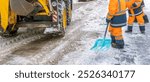 Two men in orange and blue work together to clear snow from a road. One of them is using a snow shovel while the other is using a snow plow. The scene is set in a snowy environment