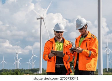 Two men in orange jackets are looking at a tablet. They are wearing hard hats. Technician team in high visibility jackets carrying toolboxes for maintenance service at renewable energy site.