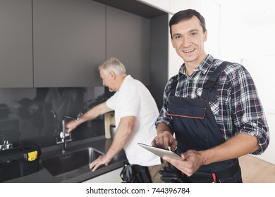 Two Men Of Plumbers Are Standing In The Kitchen. An Elderly Man Inspects The Place Of Repair Work. The Second Man Makes Notes On His Form. Next To Them Is A Black Tool Box.