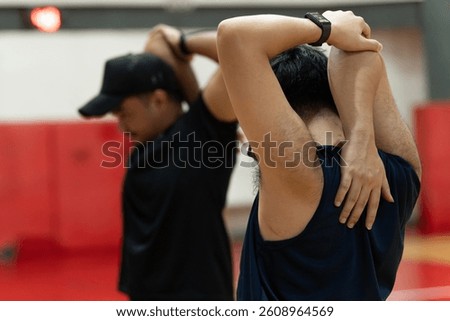 Two men raise one arm overhead and bend it behind their head while using the other hand to press the elbow, performing a triceps stretch as a warm-up before their game.