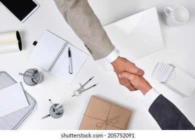 Two Men Reaching Over A Desk To Shake Hands. Shot From A High Angle The Desk Is White As Are Most Of The Office Items Except For A Brown Wrapped Package. 