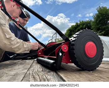Two men repairing or assembling a manual red reel lawn mower on a wooden surface outdoors.