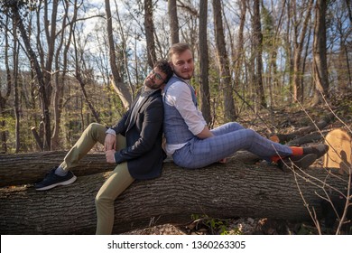 Two Men, Romantic Lovely Gay Couple, Leaning Back On Back, Posing Looking To Camera Carefree. Sitting On Log In Woods.