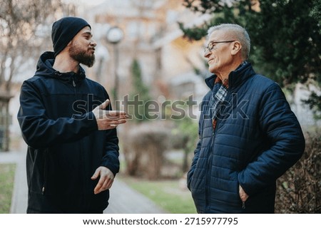 Two men share a warm chat in a park as they stand on a path, wearing jackets. The younger man gestures with his hand while the older man smiles, enjoying a friendly moment outdoors.