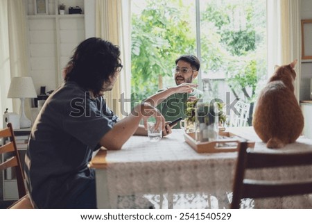 Two men sit at a dining table in a cozy, sunlit kitchen with vintage decor, engaged in conversation while a cat sits nearby, creating a relaxed setting.
