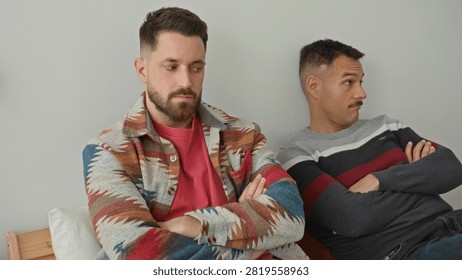 Two men sit on bed with arms crossed and turned away inside a building, one looking annoyed and the other sullen; tension distance.