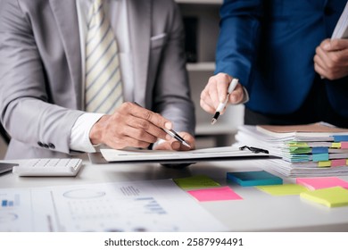 Two men are sitting at a desk with a white board and a stack of papers. One of the men is pointing at the white board while the other man looks on. The white board has a few numbers and graphs on it
