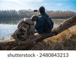 Two men sitting on a log by the river, taking photos on a smartphone, father and son, early spring landscape, the concept of hiking and outdoor activities