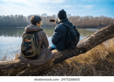 Two men sitting on a log by the river, taking photos on a smartphone, father and son, early spring landscape, the concept of hiking and outdoor activities - Powered by Shutterstock