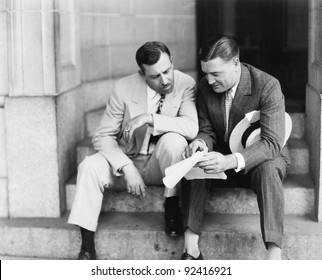 Two men sitting on steps and reading a document