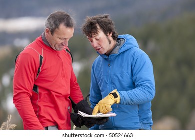 Two men stand and look down at a map together in the wilderness. One man is pointing at a spot on the map and talking. Horizontal format.