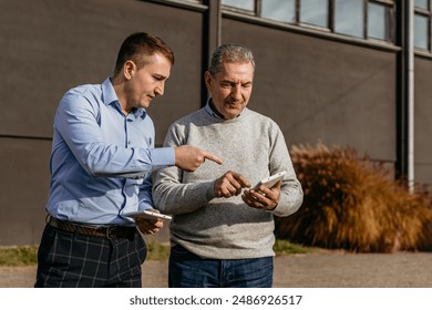 Two men stand outside a building on a sunny day. The younger man is pointing at his phone as the older man looks at the screen. The younger man is wearing a blue shirt and plaid pants.  - Powered by Shutterstock