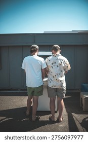 Two men stand side-by-side, facing a surface on a bright, sunny day, possibly reading or working together