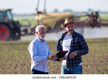 Two Men Standing In Field Looking Watching. Farmer And Insurance Sales Rep Representative Surveying In Front Of Tractor Machinery Boom Sprayer.