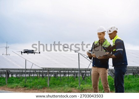 Two men are standing in a field, looking at a drone. They are wearing hard hats and are looking at a laptop. The drone is flying above them, and they seem to be controlling it