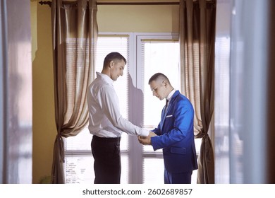 Two men are standing in front of a window, one of them is wearing a blue suit. The man in the blue suit is adjusting his tie while the other man looks on. Scene is one of formality and professionalism