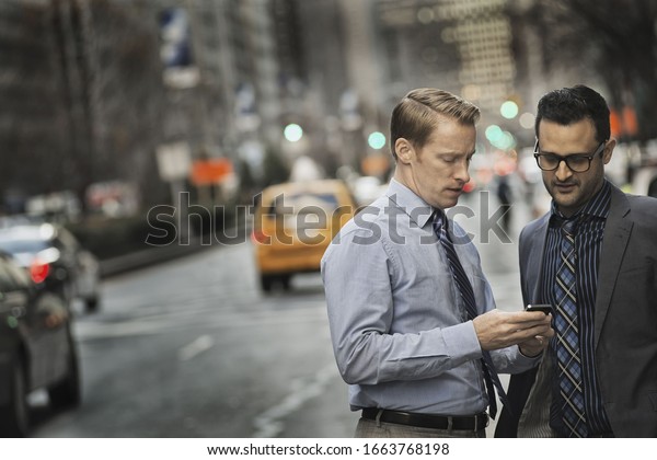 Two men standing together looking at a\
cell phone display on a busy street at\
dusk.