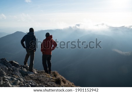 Two men standing standing with trekking poles on cliff edge and looking at sunset rays over the clouds. Successful summit concept image.