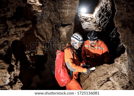 Two men, strong physique, explore the cave. Men dressed in special clothes to pass through the cave and stopped, looking at the map.