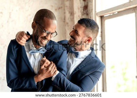 Two men in suits, smiling and embracing near a window. Both wear bow ties, creating a joyful and celebratory atmosphere.