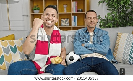 Two men supporting soccer team sitting on sofa at home