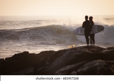 Two Men With Surf Standing At The Anchor Point In Morocco Waiting For A Wave During Sunset.