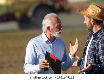 Two Men Talking Arguing Explaining In Field With Machinery In Background. Farmer And Insurance Sales Rep Representative Negotiating Holding Clipboard.