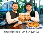 Two men in traditional Bavarian Beer garden or oktoberfest toasting with beer mugs at an table with a pretzel in Bavaria, Germany