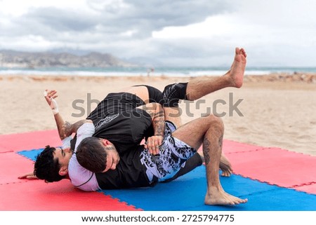 Two men training jiu jitsu on wrestling mats at the beach with mountains and sea in the background. Practicing martial arts outdoors