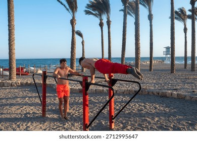Two men training on parallel bars by the sea under palm trees, building strength, balance and core fitness in a sunny outdoor workout