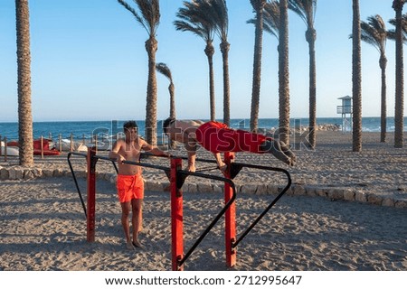 Two men training on parallel bars by the sea under palm trees, building strength, balance and core fitness in a sunny outdoor workout