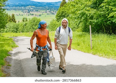 Two men walking paved path green forest, older man stepping orange tank top, younger man backpack white hoodie, Senior fitness and outdoor activity, nature trek, physical activity fatigue long trek