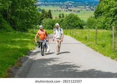 Two men walking paved path green forest, older man stepping orange tank top, younger man backpack white hoodie, Senior fitness and outdoor activity, nature trek, physical activity fatigue long trek