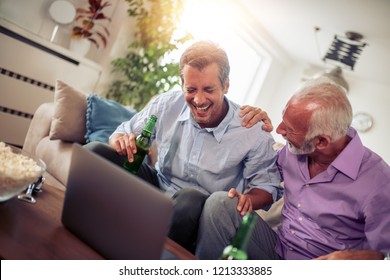 Two men watching football game,drinking beer and eating popcorn at home.
