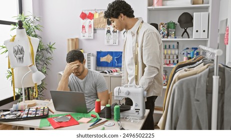 Two men working in a tailor shop, one sewing and the other consulting a laptop with a concerned expression. - Powered by Shutterstock