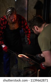 Two men in workwear processing molten metal workpiece with handtools on anvil