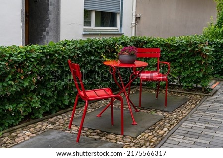 Two metal chairs and a table painted with red paint in the courtyard of the house. Surrounded by green vegetation.