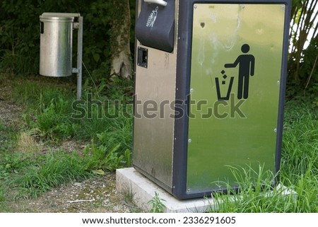 Two metal litter bins in public spaces. The first one is cuboidal and bears a icon instructing to put litter in it. The second one on the background is cylindrical. 