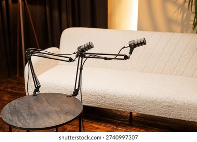 Two microphone stands are set up on a table in a studio. A light-colored couch is visible in the background. This is a place for podcast recording sessions.