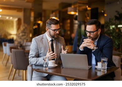 Two mid adult Caucasian male executives in suits discuss strategies over a laptop in a contemporary cafe setting, a key example of mobile office flexibility. - Powered by Shutterstock