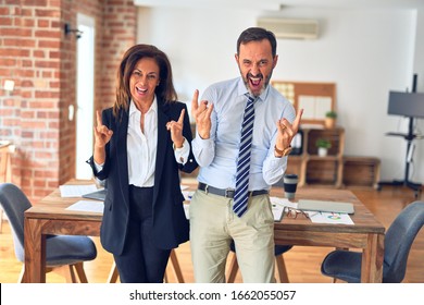 Two Middle Age Business Workers Standing Working Together In A Meeting At The Office Shouting With Crazy Expression Doing Rock Symbol With Hands Up. Music Star. Heavy Concept.