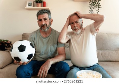 Two middle aged men expressing sadness, frustration, and disappointment while watching a football match on television at home